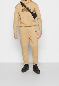 Conjunto de sudadera con capucha y pantalones deportivos beige de Nike con detalles de logo en negro. Incluye un bolsillo canguro, capucha con cordón y puños acanalados. Combinado con zapatillas negras.