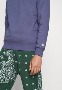 Sudadera de color púrpura oscuro con puños y dobladillo acanalados, que presenta un pequeño logo dorado. Combinada con pantalones verdes que exhiben un patrón de paisley blanco.
