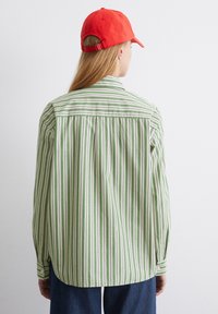 Camicia a righe verde e bianche con colletto a punta, polsini abbottonati e dettagli plissettati sul retro, abbinata a un cappellino rosso.