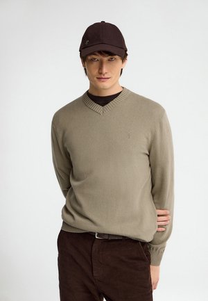 Joven con un suéter beige de cuello en V, gorra marrón oscuro y pantalones marrón oscuro, de pie con una mano detrás de la espalda.