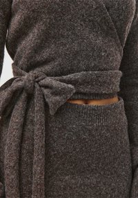 Nærbillede af en mørkebrun strikket sweater, der er bundet i en sløjfe i taljen over matchende højtaljede bukser, der viser en lille hudkløft.