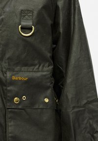 Must veekindel jope, millel on kuldne metallist D-rõngas, tikitud "Barbour" logo ja kuldsed kinnitusnupud tasku piirkonnas.