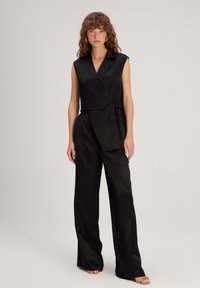 Zwarte, mouwloze jumpsuit met een wikkeldesign, voorzien van een gestrikte taille en wijde broekspijpen. Gemaakt van soepel, gestructureerd materiaal.