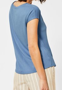 Nainen, joka pitää yshort-sleeve sinistä ykköstä ja vaaleita housuja, joissa on valkoiset pystysuorat raidat, näkyy selkä- ja sivuprofiilista yksinkertaista valkoista taustaa vasten.