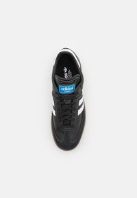 Svart lädersneaker med vita ränder och en blå logotypetikett, med rundad tå, texturerad gummisål och snörningsdesign.