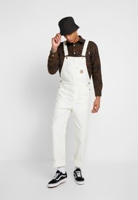 Salopette blanche en coton avec bretelles réglables, quincaillerie dorée et poche avant. Associée à une chemise à motifs marron et des baskets noires.