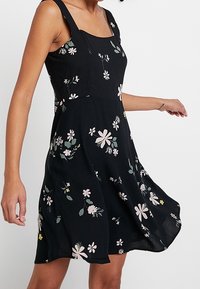 Robe noire à fleurs avec un décolleté carré, des bretelles épaisses et une jupe évasée. Présente diverses fleurs aux couleurs pastel et des feuilles vertes.