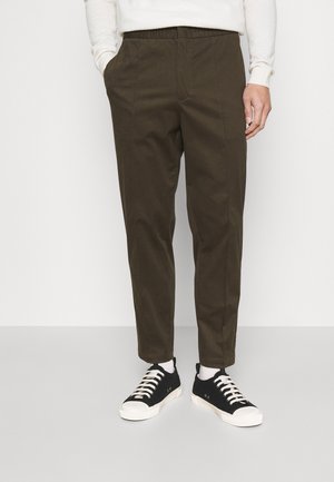 Pantalons kaki avec une taille élastique, poches latérales, coupe droite et détails de couture minimaux, associés à des baskets noires.