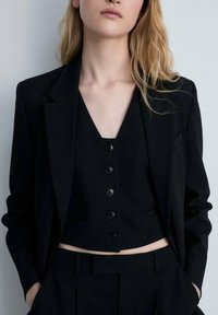 Černý dobře střižený blazer přes sladěný bezrukávový top s výstřihem do V a pěti knoflíkovými zapínáními, doplněný o úzké černé kalhoty.