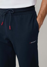 Marinefarbene Jogginghosen aus weichem Material mit elastischem Bund, Kordelzug mit roten Spitzen und einem weißen und roten Logotext auf dem Oberschenkel.