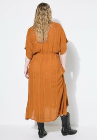 Rostfarbene Maxikleid mit strukturierten vertikalen Streifen und Taschen, mit einem lockeren Schnitt und kurzen Dolmanärmeln. Getragen mit genagelten schwarzen Stiefeln.