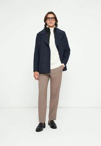 Uomo che indossa un cappotto nero, un maglione bianco a collo alto, pantaloni beige, mocassini neri e occhiali, in piedi davanti a uno sfondo bianco.