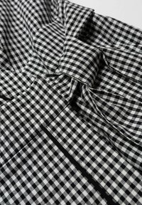 Czarna i biała tkanina w kratkę o fakturowanym splocie, z fałdami i detalem w postaci kokardy, podkreślona kontrastującym czarnym ściegiem.