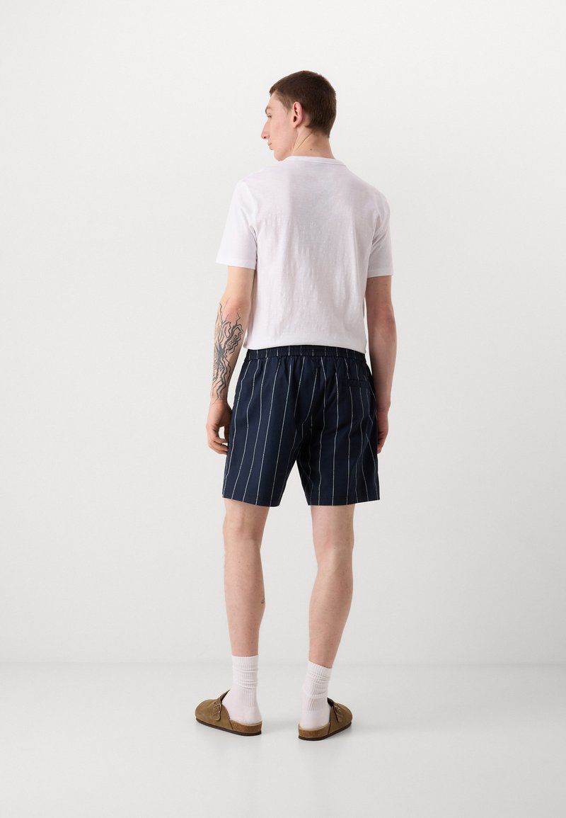 Marineblau gestreifte Shorts mit einem elastischen Bund, kombiniert mit einem simplen weißen T-Shirt. Das Outfit wird durch braune Slipper und weiße Socken ergänzt.