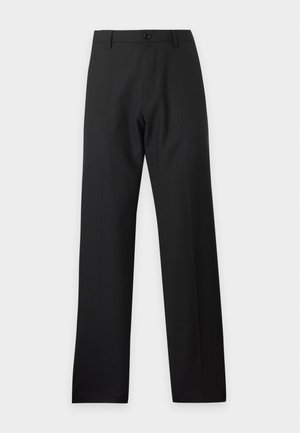 Jeune homme portant une chemise à manches longues rayée verte et blanche, pantalon noir et baskets noires, debout devant un fond blanc uni.
