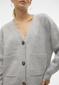 Cardigan gris clair en tissu doux, avec un col en V, trois grands boutons, des poignets côtelés et deux poches avant.