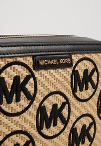 Vävd halmhandväska med svarta läderdetaljer, guldfärgade metalldelar och ett upprepat svart MK-logomönster på en beige bakgrund.