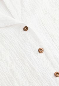 Tejido blanco texturizado con un patrón de diamantes y tres botones marrones de cuatro agujeros alineados verticalmente en la prenda.