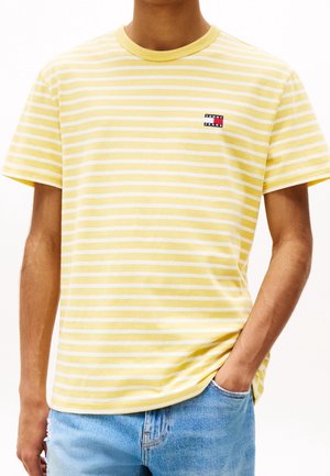 Mężczyzna w żółto-białej, pasiastiej koszulce z krótkim rękawem z logo Tommy Jeans, noszący jasnoniebieskie dżinsy, jedną ręką w kieszeni.