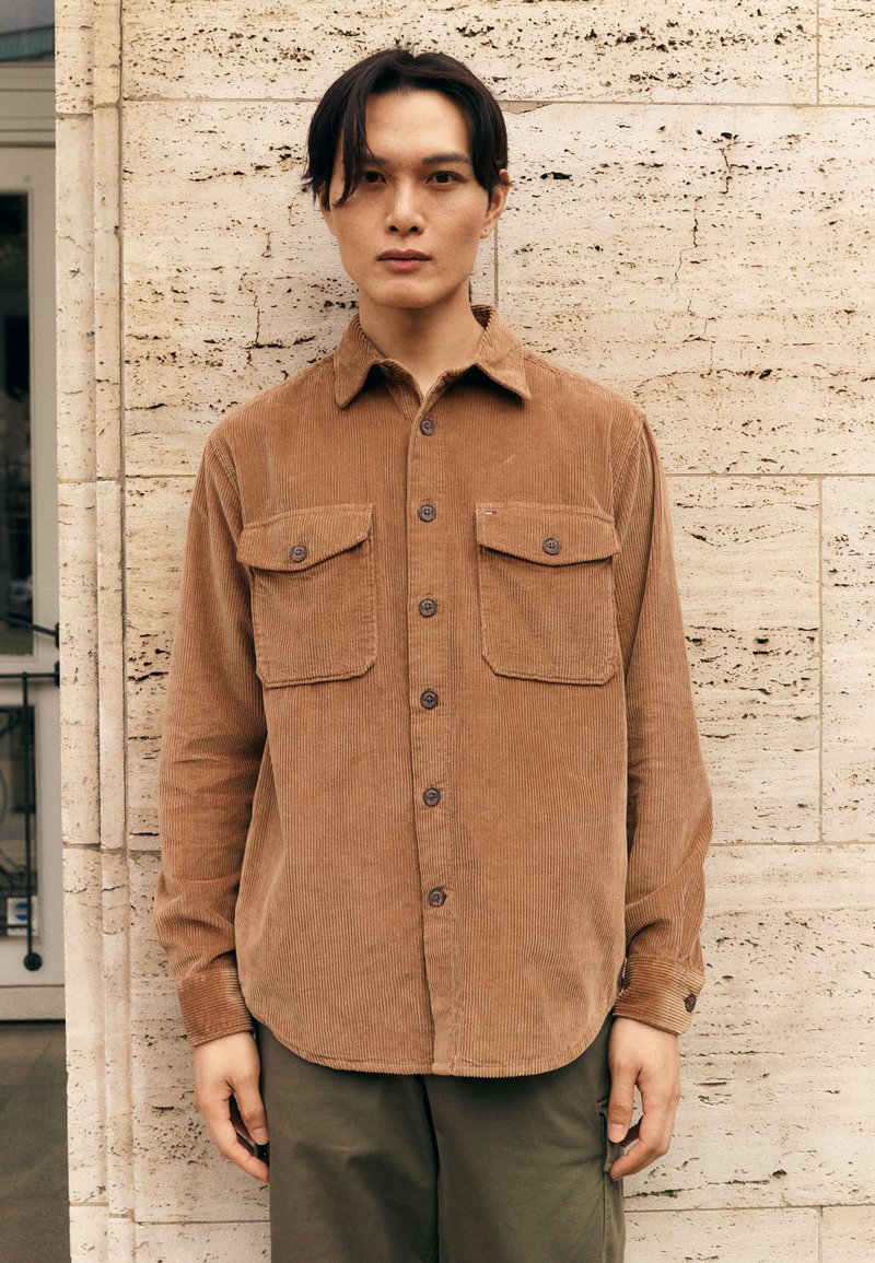 Brown corduroy shirt with two chest pockets, button front, and long sleeves. The fabric has a ribbed texture. Worn over olive green pants.