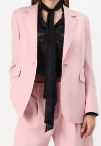 Blazer rose clair sur mesure avec un seul bouton à l'avant, associé à un chemisier transparent noir à fleurs et une écharpe noire, présentant une coupe décontractée.