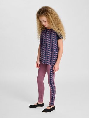 Jong meisje met krullend blond haar dat een patroonrijke marineblauwe en roze kortemouwhemd en bijpassende roze legging met marineblauwe details draagt, staand.
