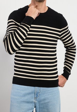 Homme portant un pull à rayures horizontales noires et blanches et un jean bleu clair, une main près du cou, l’autre sur la hanche, sur fond blanc.