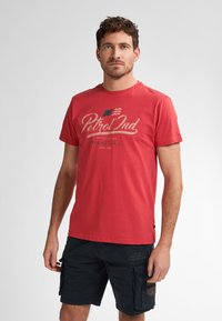 Camiseta de algodón roja con mangas cortas, cuello redondo y gráficos en la parte frontal. Se lleva con pantalones cortos cargo oscuros que tienen bolsillos laterales y detalles.