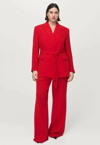 Woman wearing a bold red tailored suit with a belted blazer and wide-leg trousers, standing against a plain light background.