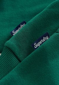 Zelená mikina so širokými manžetami. Obsahuje modrú látkovú nálepku s bielym nápisom "Superdry". Mäkká textúra a ležérny dizajn.