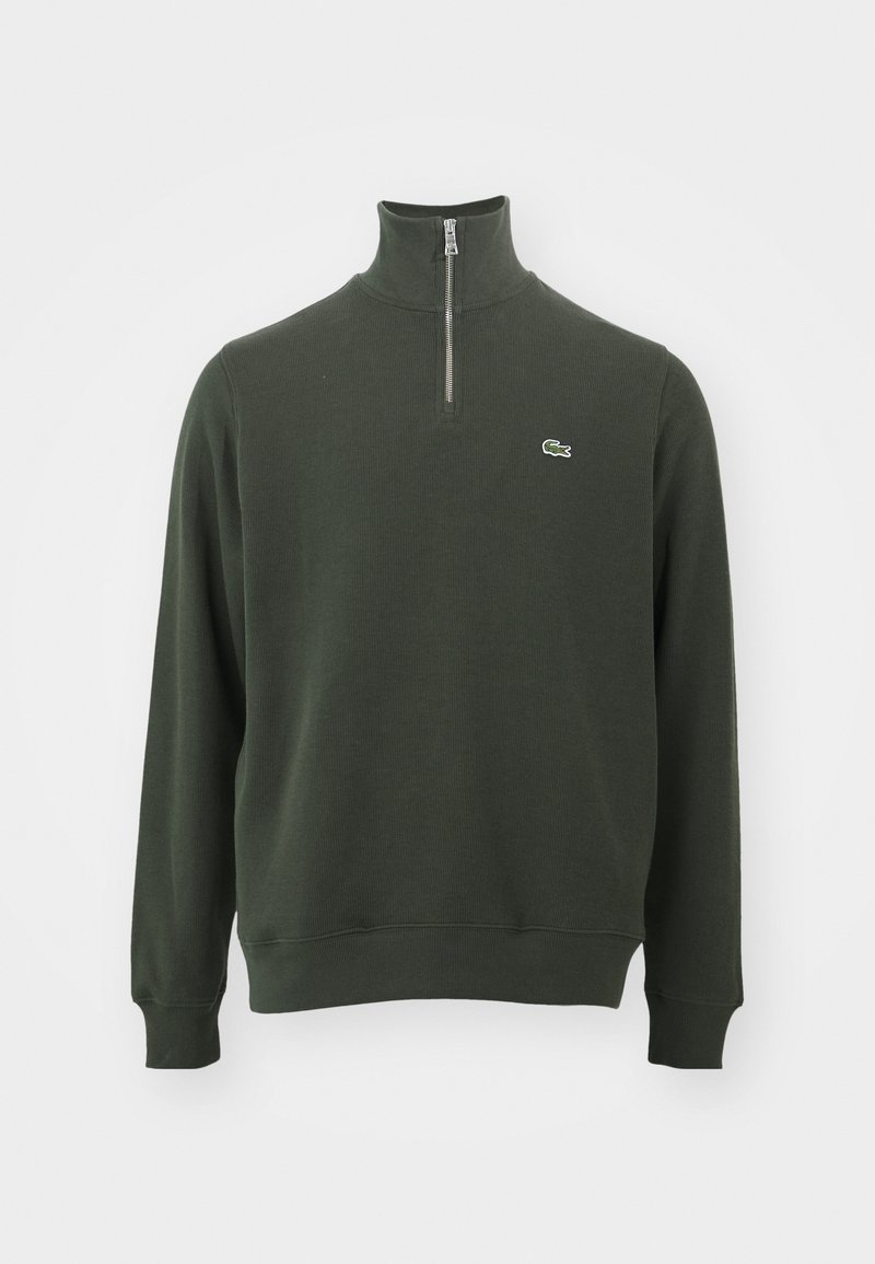 Pull en polaire vert foncé à manches longues et zip quart, avec une texture côtelée et un petit logo de crocodile brodé sur la poitrine, se tenant devant un fond blanc.
