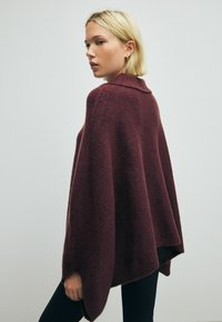 Bordeaux strikket poncho med krave, løs pasform og blød tekstur, der hænger over skuldrene og strækker sig til hoften.
