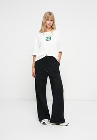 Camiseta de algodón blanca con gráfico del número "23" en verde, de corte holgado, combinada con pantalones de chándal negros de pierna ancha con cordón ajustable.