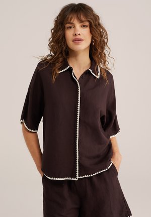 Frau mit lockigem Haar, die ein dunkelbraunes kurzärmeliges Shirt und passende Shorts mit weißem scallopförmigem Saum trägt, Hände in den Taschen, neutraler Hintergrund.