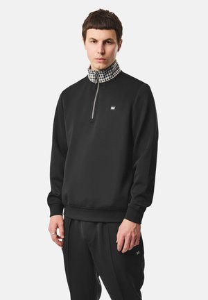 Jeune homme portant un sweat-shirt noir à demi-fermeture éclair avec col haut à carreaux et pantalon noir assorti, debout devant un fond uni.