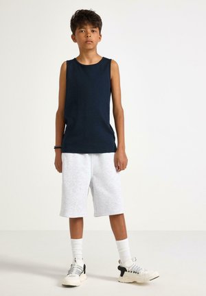 Jongen staat met een marineblauw mouwloos shirt, lichtgrijze knielange shorts, witte sokken en witte sneakers op een effen achtergrond.
