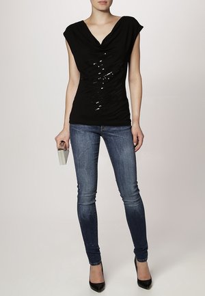 Černý top s krátkým rukávem a splývavým výstřihem s flitrovými detaily na přední straně. Kombinován s přiléhavými modrými džíny a černými lodičkami.