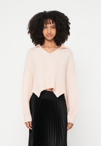 Ροζ ανοιχτόχρωμο κοντό πουλόβερ με V-λαιμόκοψη και γιακά, με χαλαρή εφαρμογή και υφή πλεκτού. Συνδυάζεται με μαύρη πλισέ φούστα.