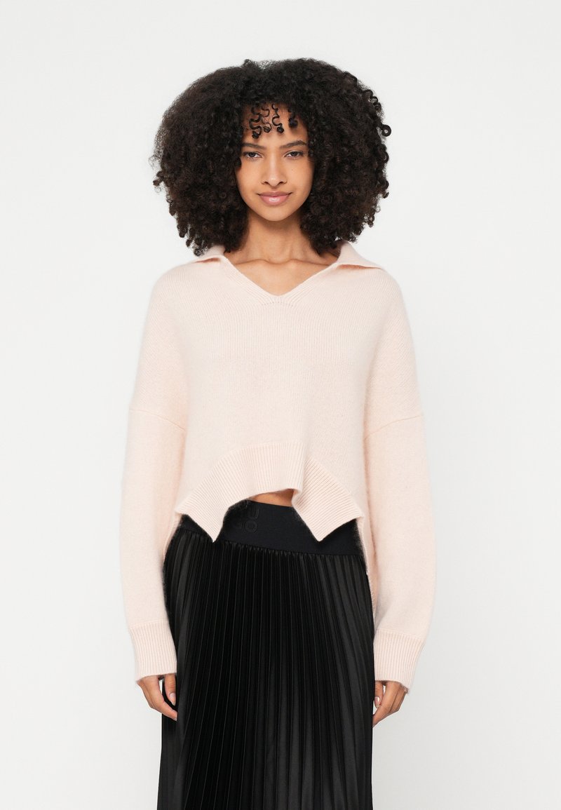 Ροζ ανοιχτόχρωμο κοντό πουλόβερ με V-λαιμόκοψη και γιακά, με χαλαρή εφαρμογή και υφή πλεκτού. Συνδυάζεται με μαύρη πλισέ φούστα.