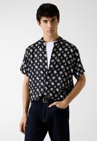 Chemise noire à manches courtes avec un motif géométrique blanc, col et fermeture à boutons, associée à un jean foncé et une ceinture noire.