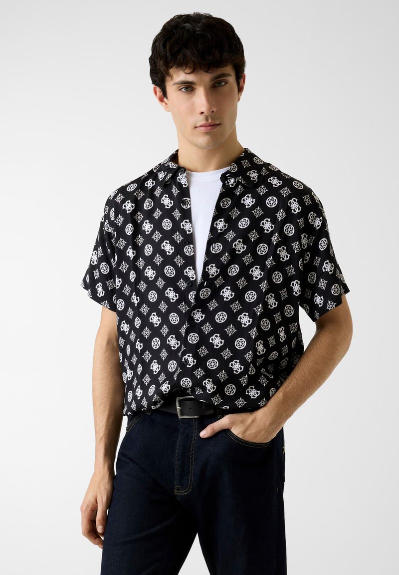 Chemise noire à manches courtes avec un motif géométrique blanc, col et fermeture à boutons, associée à un jean foncé et une ceinture noire.
