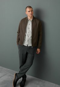 Cazadora bomber marrón oscuro, camiseta de botones de patrón gris claro, pantalones grises y zapatillas negras con suelas de goma, sobre un fondo gris.