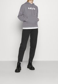 Sudadera gris con el logo blanco de "Levi's", bolsillo lateral, llevada sobre una camiseta blanca, combinada con jeans rectos negros y botas negras gruesas.