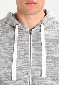 Sweat à capuche zippé gris avec un tissu texturé, des cordons blancs et une fermeture éclair argentée. Le design offre une coupe décontractée et confortable.