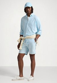 Ανοιχτό μπλε πουκάμισο με κουμπιά, ταιριαστά σορτς και καπέλο· απαλό ύφασμα με άνετη εφαρμογή· πουλόβερ δεμένο γύρω από τη μέση· λευκά αθλητικά παπούτσια.