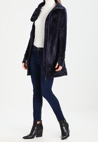 Donna che indossa una giacca a zip blu navy, un dolcevita bianco, jeans skinny scuri e stivaletti neri con tacco, in piedi contro uno sfondo bianco.
