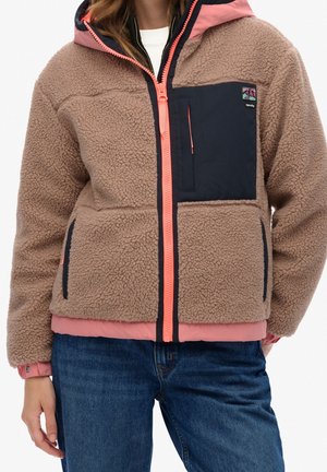 Persona que lleva una chaqueta de forro polar color beige con cremallera coral y bolsillo en el pecho color azul marino, combinada con vaqueros azules.