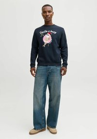 Navy sweatshirt met de tekst "Santa is busy" en een afbeelding van een kerstman in een opblaasband. Gecombineerd met lichtblauwe wijde jeans en beige schoenen.