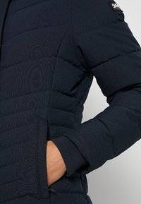 Giacca imbottita blu navy con texture trapuntata, dotata di tasca con cerniera e polsini elasticizzati. Realizzata in tessuto sintetico per calore e comfort.