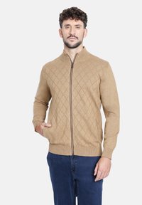 Cárdigan de cremallera en tono beige con un patrón de diamantes, puños y dobladillo acanalados, y dos bolsillos frontales. Fabricado con un material suave y texturizado.
